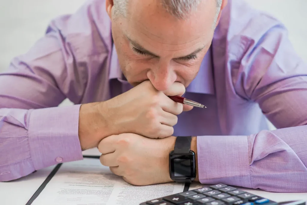 man looking depressed with calculator beside him and looking down at documents on a table